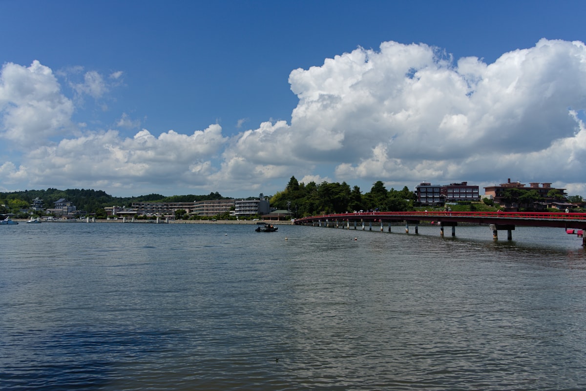 A red bridge crosses a calm bay with buildings beyond.