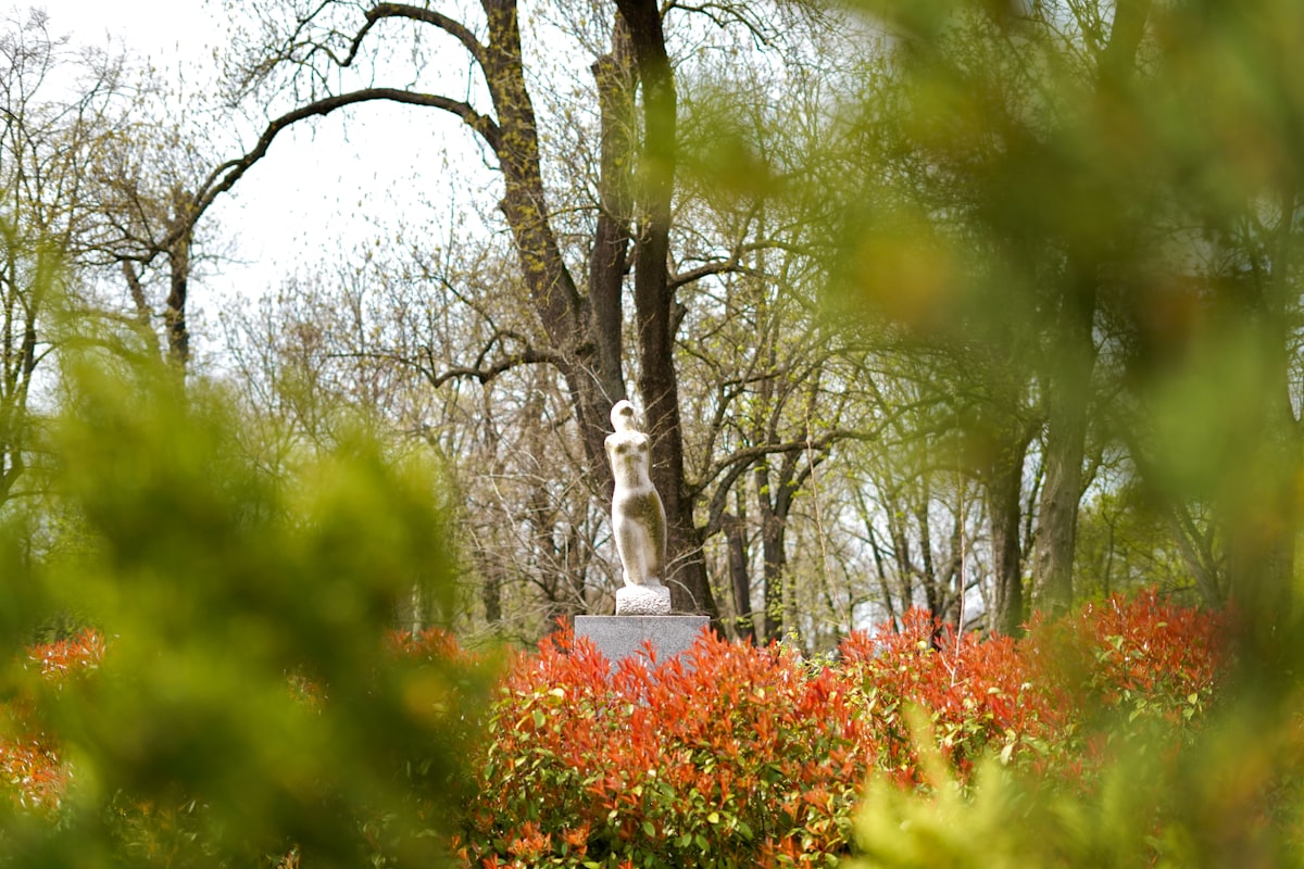 A sculpture stands in a park with trees and bushes.
